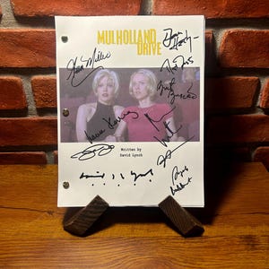 Peut inclure: Un scénario signé du film "Mulholland Drive" avec une photo de deux femmes. Le script est soutenu par deux petits supports en bois foncé. Le titre et les signatures sont à l'encre noire.
