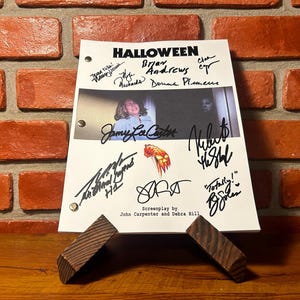 Può includere: Una pagina di sceneggiatura bianca per il film "Halloween" con firme e il titolo del film in alto. La sceneggiatura è appoggiata su un supporto di legno contro un muro di mattoni.