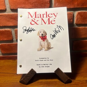 May include: A script for the movie "Marley & Me" featuring the title in red, a puppy wearing a red bow, and signatures. The script is held by two wooden supports against a brick wall.