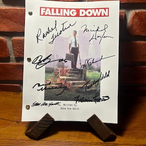 May include: A signed script for the movie "Falling Down" is displayed on a wooden surface. The script features the title in red, along with several signatures. The image includes a photo of a man holding a briefcase.