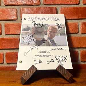 Könnte beinhalten: Ein Drehbuch für den Film "Memento" mit dem Titel und Unterschriften. Das Skript wird von zwei kleinen Holzblöcken getragen. Der Hintergrund ist eine rote Backsteinmauer.