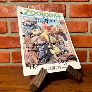 Könnte beinhalten: Ein weißes Papier mit dem Titel "Zootopia" und Charakterillustrationen, signiert von mehreren Personen. Das Papier steht auf einem Holzständer vor einer Backsteinmauer. Das Drehbuch stammt von Jared Bush und Phil Johnston.
