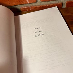 Puede incluir: Libro abierto con el t&iacute;tulo "CRY-BABY" de John Waters, Tercera versi&oacute;n, 22 de febrero de 1989. Las p&aacute;ginas son blancas con texto negro. El fondo incluye una pared de ladrillo y una superficie de madera.