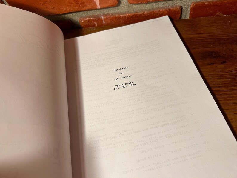 Puede incluir: Libro abierto con el t&iacute;tulo "CRY-BABY" de John Waters, Tercera versi&oacute;n, 22 de febrero de 1989. Las p&aacute;ginas son blancas con texto negro. El fondo incluye una pared de ladrillo y una superficie de madera.