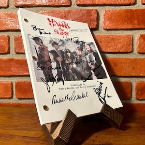 Op de afbeelding: Een gesigneerd script voor "Hawk the Slayer" op een houten standaard. Het script bevat een castfoto en meerdere handtekeningen. De achtergrond is een bakstenen muur.