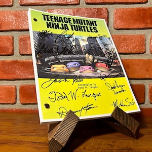 May include: A yellow script for "Teenage Mutant Ninja Turtles" with multiple signatures. The script features an image of the turtles and is propped up on a wooden stand against a brick wall.