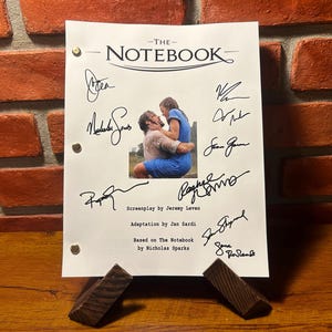 Może przedstawiać: Biały scenariusz do filmu "The Notebook" z podpisami obsady i ekipy. Scenariusz stoi na drewnianym stojaku i zawiera czarno-białe zdjęcie głównych bohaterów.
