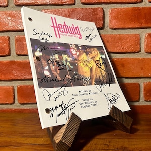 May include: A script for "Hedwig and the Angry Inch" on a wooden stand. The white script has the title in pink and black, a photo, and multiple signatures. It is propped up on a wooden stand against a brick wall.