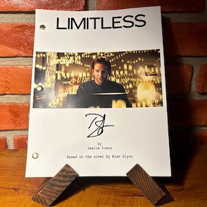 Peut inclure: Un script blanc avec le titre "LIMITLESS" en haut, avec une photo d'un homme. Le script est soutenu par deux supports en bois. Le script est basé sur le roman d'Alan Glynn.