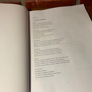 May include: Open book with text from Act 1 of the musical "Hamilton." The page features the title "Alexander Hamilton" and lines of dialogue from the play. The text is printed in black ink on white paper.