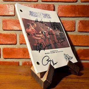 Puede incluir: Un guion firmado de "Jersey Boys" en un soporte de madera. El guion presenta una foto del elenco con trajes rojos. El fondo es una pared de ladrillo y una superficie de madera.