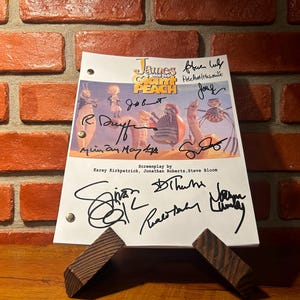 May include: A script from the movie "James and the Giant Peach" with multiple signatures. The script is propped up on a wooden stand in front of a brick wall. The title is in yellow and the script is in English.