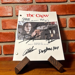 Puede incluir: Un guion blanco con el título "The Crow" en rojo, con una foto de un hombre sosteniendo un arma y varias firmas. El guion está sostenido por un soporte de madera contra una pared de ladrillos.