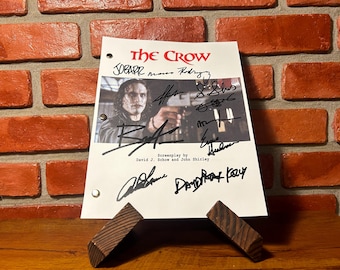 May include: A white script with the title "The Crow" in red, featuring a photo of a man holding a gun, and multiple signatures. The script is propped up by a wooden stand against a brick wall.