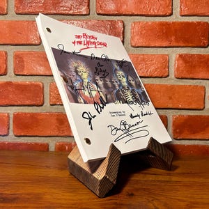 May include: A script for "The Return of the Living Dead" rests on a wooden stand. The script is white with black text and signatures. The cover features a zombie-themed illustration. The stand is made of dark wood.