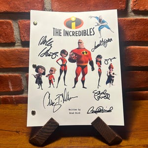 May include: A white paper featuring the title "The Incredibles" with character illustrations and signatures. The artwork includes the main characters in their superhero costumes. The paper is propped up by two wooden supports against a brick wall.