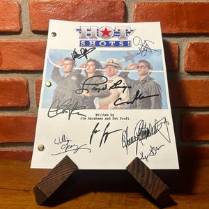 Op de afbeelding: Een gesigneerd script van de film "Hot Shots!" met een foto van de cast en meerdere handtekeningen. Het script staat op een houten standaard tegen een bakstenen muur.