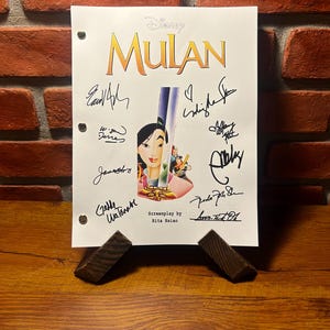 May include: A white sheet of paper with the Disney film title "Mulan" in gold lettering, featuring a central illustration of the character Mulan. The sheet is signed by multiple individuals and is propped up by two small, dark brown wooden supports.