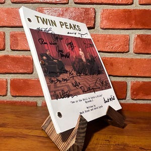 May include: A script for the television show "Twin Peaks" with multiple signatures, displayed on a wooden stand. The script is titled "Twin Peaks" and includes the episode title "Ian or the Skill to Catch a Killer". The background is a red brick wall.