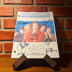 Puede incluir: Un guion firmado de la película "Anchorman: The Legend of Ron Burgundy" con el elenco. El guion se muestra en un soporte de madera contra un fondo de pared de ladrillo. El título "ANCHORMAN" es visible en la parte superior, junto con los nombres de los actores.