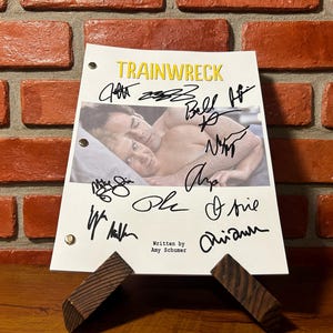 May include: A script for the movie "Trainwreck" with signatures, displayed on a wooden stand. The script has a photo of two people and the title "Trainwreck" in yellow. The background is a brick wall.