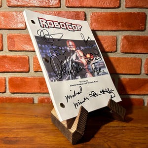 Puede incluir: Un guion de la película Robocop, apoyado en un soporte de madera, con múltiples firmas. El guion presenta el logotipo de Robocop y una imagen del personaje. El fondo es una pared de ladrillo.