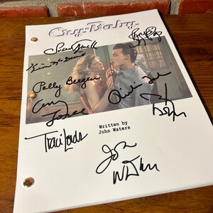 Puede incluir: Un guion blanco de la pel&iacute;cula "Cry-Baby" con m&uacute;ltiples firmas en tinta negra. El guion presenta una foto de dos actores y el texto "Written by John Waters". El guion est&aacute; sobre una superficie de madera.