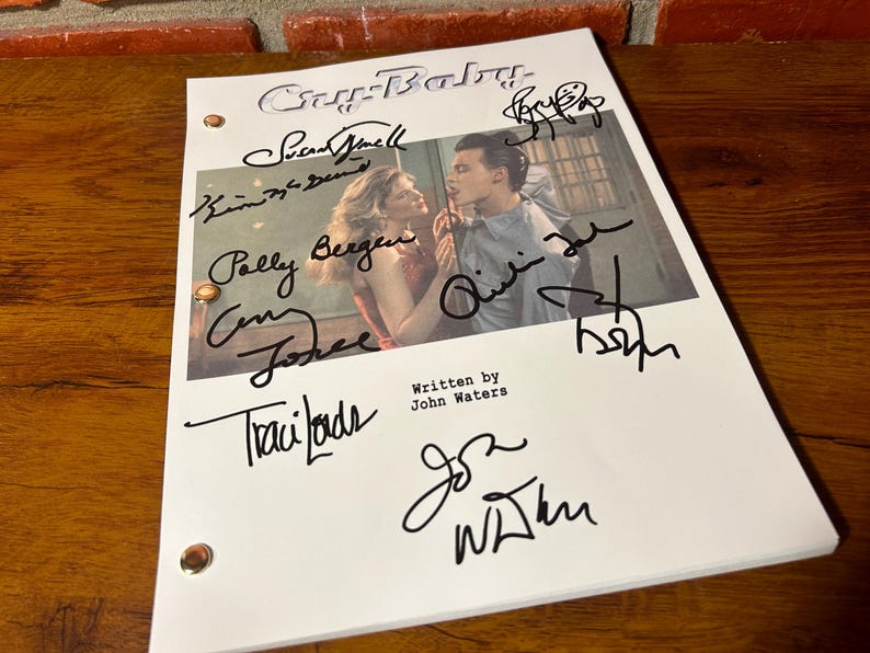 Puede incluir: Un guion blanco de la pel&iacute;cula "Cry-Baby" con m&uacute;ltiples firmas en tinta negra. El guion presenta una foto de dos actores y el texto "Written by John Waters". El guion est&aacute; sobre una superficie de madera.