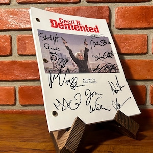 May include: A white script for the film "Cecil B. Demented" with multiple signatures. The script is propped up on a wooden stand against a brick wall. The film's title is in red, with a photo of a person holding guns.