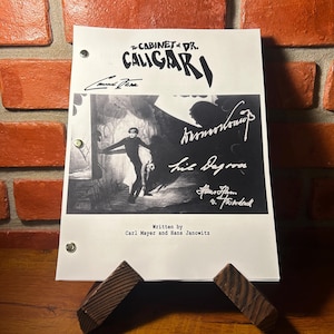 May include: A black and white print of "The Cabinet of Dr. Caligari" film script, featuring a dramatic illustration of a figure. The script is held by a wooden stand against a brick wall. Signatures and text are visible.