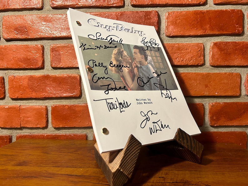 Puede incluir: Un guion blanco con firmas y una foto de una pareja bes&aacute;ndose, apoyado en un soporte de madera. El guion se titula "Cry-Baby" y est&aacute; escrito por John Waters. El fondo es una pared de ladrillos rojos.