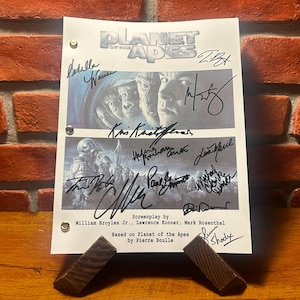 May include: A signed Planet of the Apes film script, displayed on a wooden stand against a brick wall. The script features the film's title and several signatures. The script is held in place by two small wooden stands.