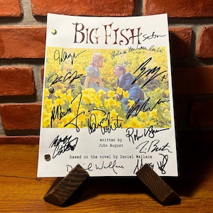 May include: A black and white photo of a couple standing in a field of yellow flowers. The photo is on a white page with black text that reads "BIG FISH", "written by John August", and "based on the novel by Daniel Wallace". The page is signed by multiple people, including the actors and the writer.