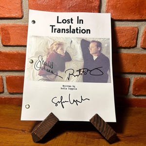 Könnte beinhalten: Ein weißes Papier mit dem Titel "Lost In Translation" und Bildern von zwei Personen, mit Unterschriften. Das Papier wird von einem Holzständer gehalten, vor einer Backsteinmauer und einer Holzoberfläche.