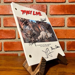 May include: A white script with the title "THEY LIVE" in red, featuring a movie still and signatures. The script is propped up on a wooden stand against a brick wall. The script is for the movie "They Live".