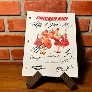 May include: A white script from the animated film "Chicken Run" featuring the film's title and character illustrations. The script is signed by multiple individuals and is propped up on a wooden stand against a brick wall.