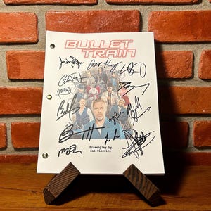 May include: A white script with the title "BULLET TRAIN" in red, featuring multiple signatures and a cast photo. The script is held by a wooden stand against a brick wall. The screenplay is by Zak Olkewicz.