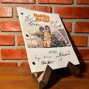 Może przedstawiać: Scenariusz filmu "Harold i Maude" z podpisami, prezentowany na drewnianym stojaku. Okładka scenariusza zawiera zdjęcie z filmu. Tło to czerwona ceglana ściana.