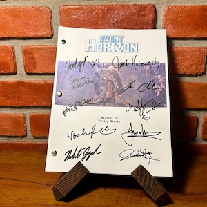 Könnte beinhalten: Ein signiertes Drehbuch für "EVENT HORIZON" auf einem Holzständer. Das Skript zeigt ein Filmbild und mehrere Unterschriften. Der Hintergrund ist eine rote Backsteinmauer.