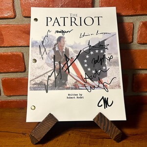 May include: A script for "The Patriot" movie, featuring the title and signatures. The script is propped up on a wooden stand against a brick wall background. The script has a picture of a man holding a flag.
