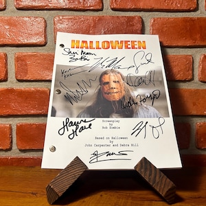 May include: A white paper with the title "HALLOWEEN" in orange and yellow, featuring a photo of a person wearing a mask. The paper is signed by multiple people and is propped up by two wooden stands. The background is a brick wall.