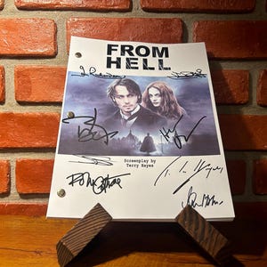 Op de afbeelding: Een script voor de film "From Hell" met handtekeningen, gepresenteerd op een houten standaard. De omslag van het script toont afbeeldingen van de acteurs en de filmtitel. De achtergrond is een bakstenen muur.