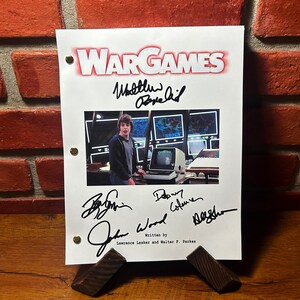 Op de afbeelding: Een wit script met de titel "WARGAMES" in rode letters. Het script is gesigneerd door Matthew Broderick, John Wood, David Gelernter en een vierde handtekening. Het script bevat een zwart-witfoto van Matthew Broderick uit de film.