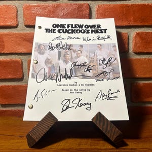 May include: A white paper with the title "ONE FLEW OVER THE CUCKOO'S NEST" and signatures, propped up by a wooden stand. The paper has a photo of men in white shirts. The background is a brick wall.