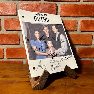 May include: A script for "American Gothic" with cast signatures, displayed on a wooden stand. The script features a cast photo and the title in bold letters. The background is a brick wall.