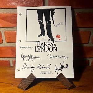 Puede incluir: Una impresión en blanco de la película "Barry Lyndon" con firmas. La impresión presenta una silueta negra de una persona con botas sosteniendo un arma, con el título y el crédito del guion. La impresión está sujeta por dos soportes de madera.