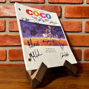 May include: A script for the Disney Pixar film "Coco" is displayed on a wooden stand. The script features the film's title and signatures. The background is a brick wall and a wooden surface.