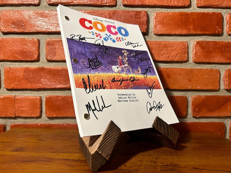 May include: A script for the Disney Pixar film "Coco" is displayed on a wooden stand. The script features the film's title and signatures. The background is a brick wall and a wooden surface.
