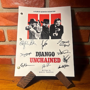 Puede incluir: Un guion de película blanco para "Django Unchained" de Quentin Tarantino, con un gráfico de tres hombres con sombreros. El título está en letras rojas grandes. El guion está sostenido por un soporte de madera y contra una pared de ladrillos.