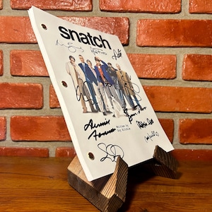 May include: A script for the movie "Snatch" with the title at the top and signatures of the cast. The script is propped up on a wooden stand against a brick wall.